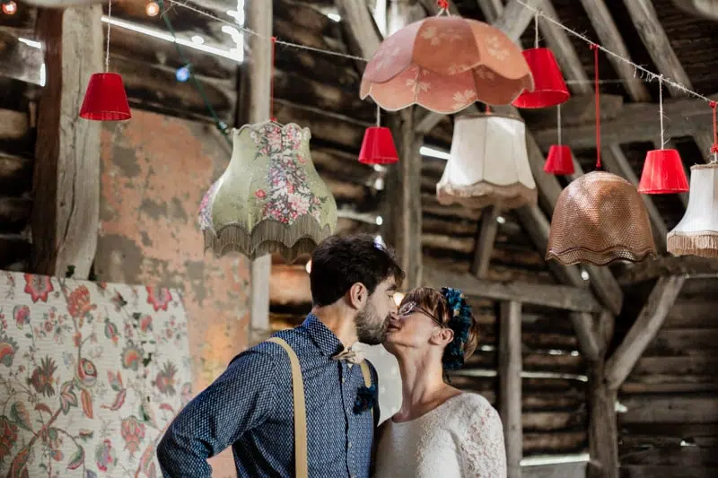 Les Marié s'embrassent avec des lustres au-dessus leur tête dans une grange en bois de style champêtre