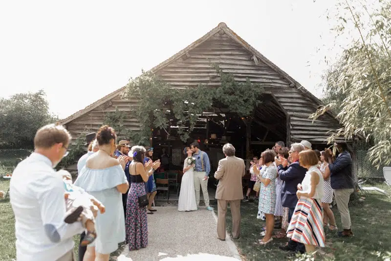 Sortie du couple de la cérémonie laïque avec les invités de chaque côté devant une grange en bois
