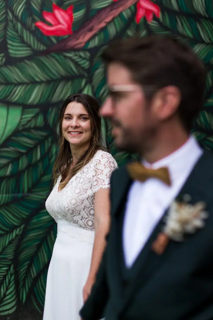 portrait de la mariée devant un mur végétale en peinture avec le marié au premier plan