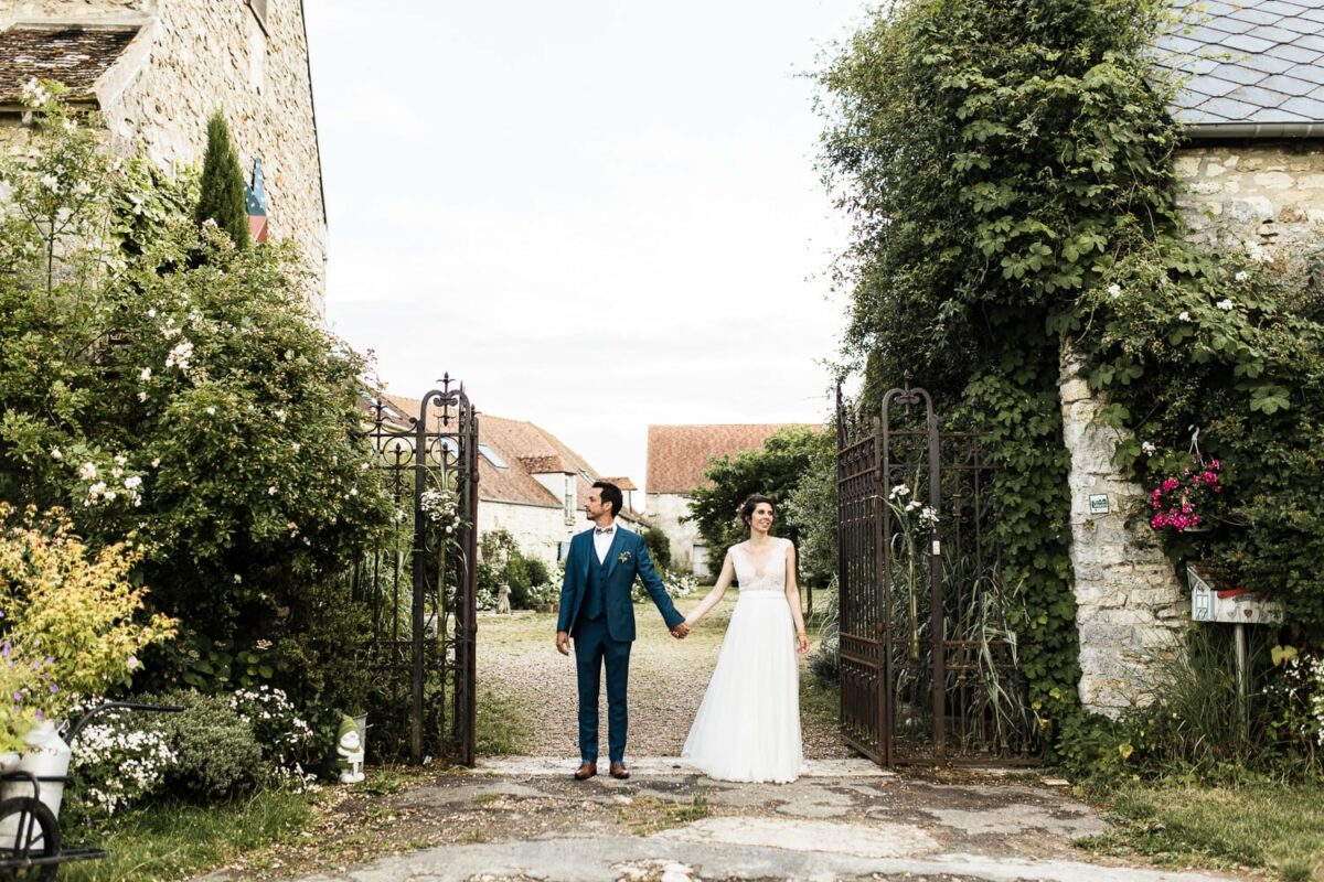 Mariés main dans la main à l’entrée de la Ferme de la Bretèche – portail et jardin champêtre.
