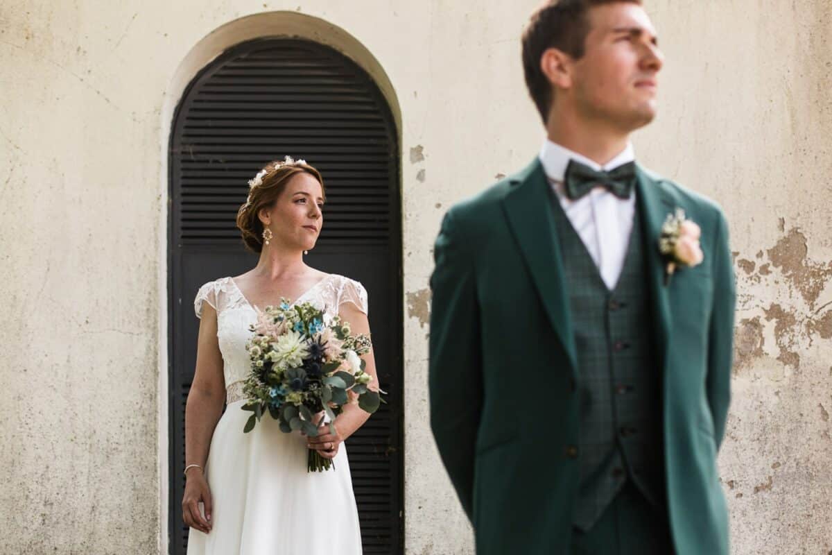 Mariée nette devant porte cintrée, bouquet en main ; marié flou au premier plan en costume vert.
