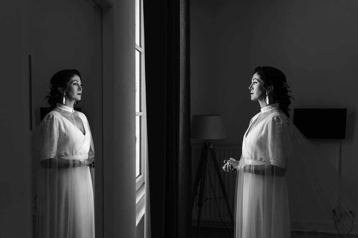 Portrait de la mariée à la fenêtre, reflet dans la glace, lumière naturelle douce.