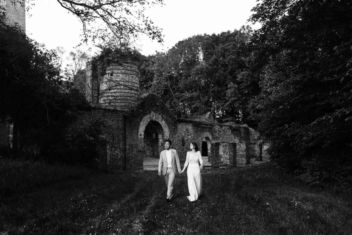 Couple de mariés marchant main dans la main devant des ruines en pierre, plan large en noir et blanc.