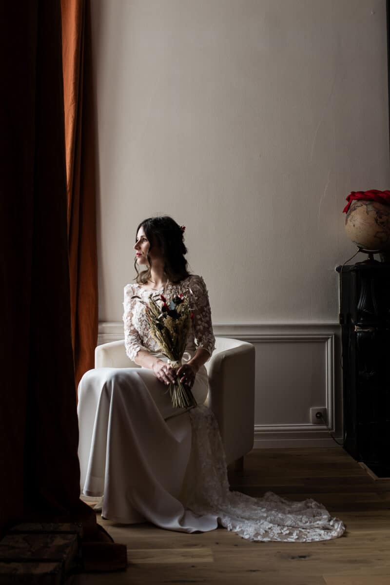 Portrait de la mariée assise près de la fenêtre, bouquet de fleurs séchées, robe en dentelle, lumière douce.