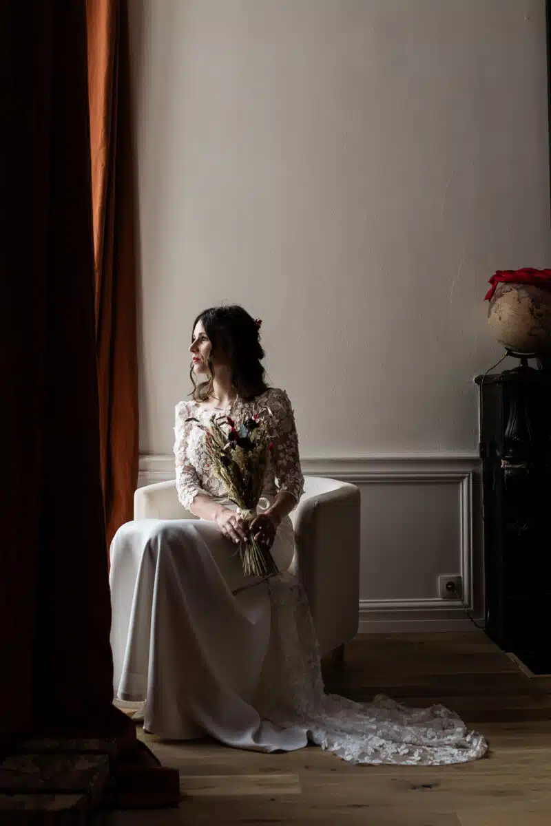 Portrait de la mariée assise près de la fenêtre, bouquet de fleurs séchées, robe en dentelle, lumière douce.