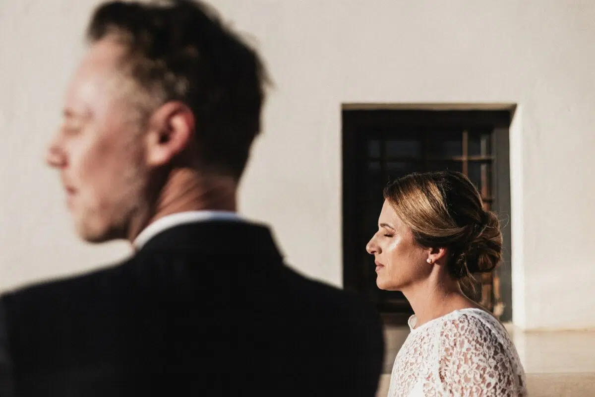 Portrait croisé des mariés en lumière dorée : mariée de profil nette, marié flou au premier plan, mur blanc et fenêtre de riad — mariage au Maroc (médina).