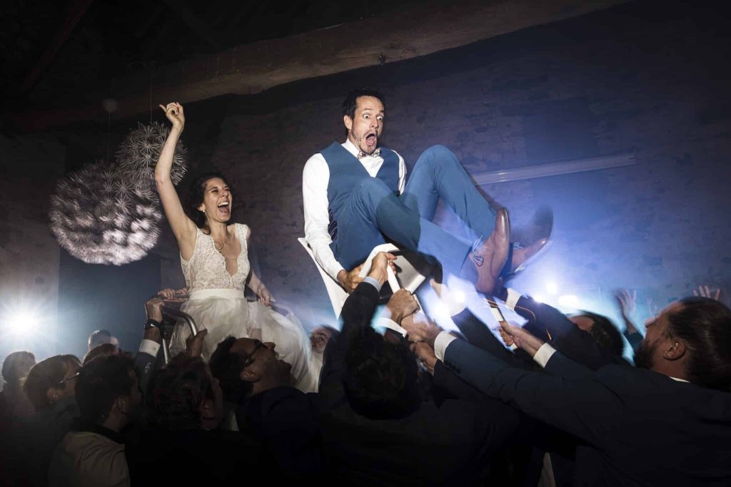 Photo de reportage de mariage à la Ferme de Genainville montrant les mariés portés sur des chaises par leurs invités dans une ambiance festive et obscure, éclairée par des projecteurs de scène.