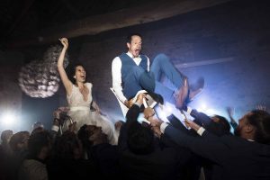 Photo de reportage de mariage à la Ferme de Genainville montrant les mariés portés sur des chaises par leurs invités dans une ambiance festive et obscure, éclairée par des projecteurs de scène.