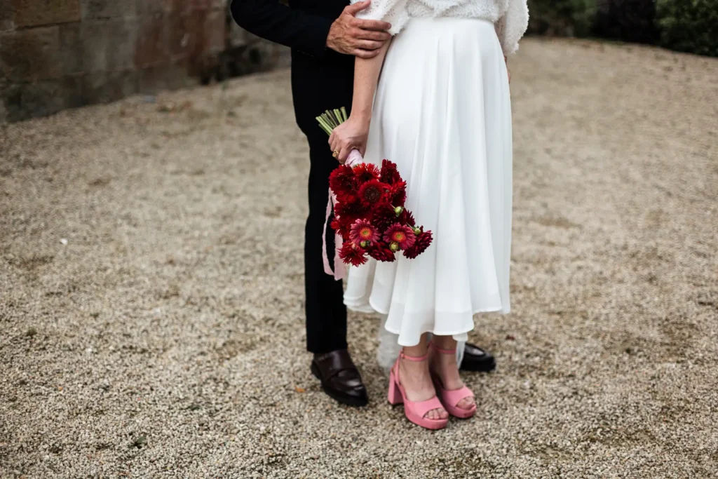 Détails de mariage : bouquet de fleurs rouges, chaussures roses de la mariée et costume du marié.