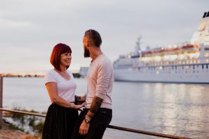 Photographie d'un couple sur les quais de Trentemoult à la tombée de la nuit, observant un immense paquebot de croisière illuminé qui remonte la Loire.