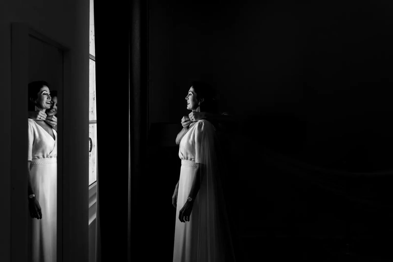 Portrait de mariage en noir et blanc : reflet de la mariée dans un miroir pendant l'ajustement de sa robe de mariée.