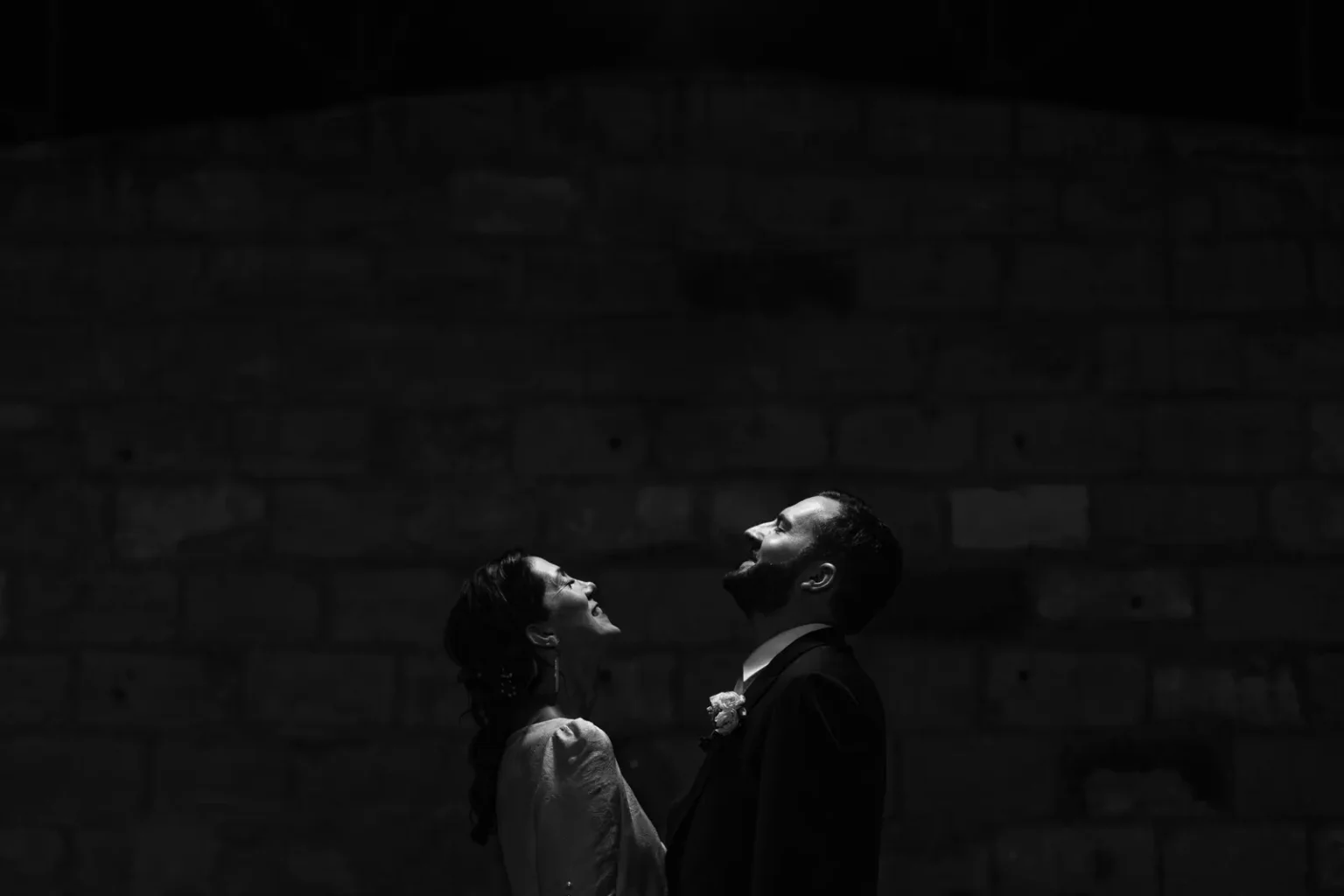 Photo de mariage artistique en clair-obscur : les mariés riant ensemble sur un fond de mur en pierre sombre.