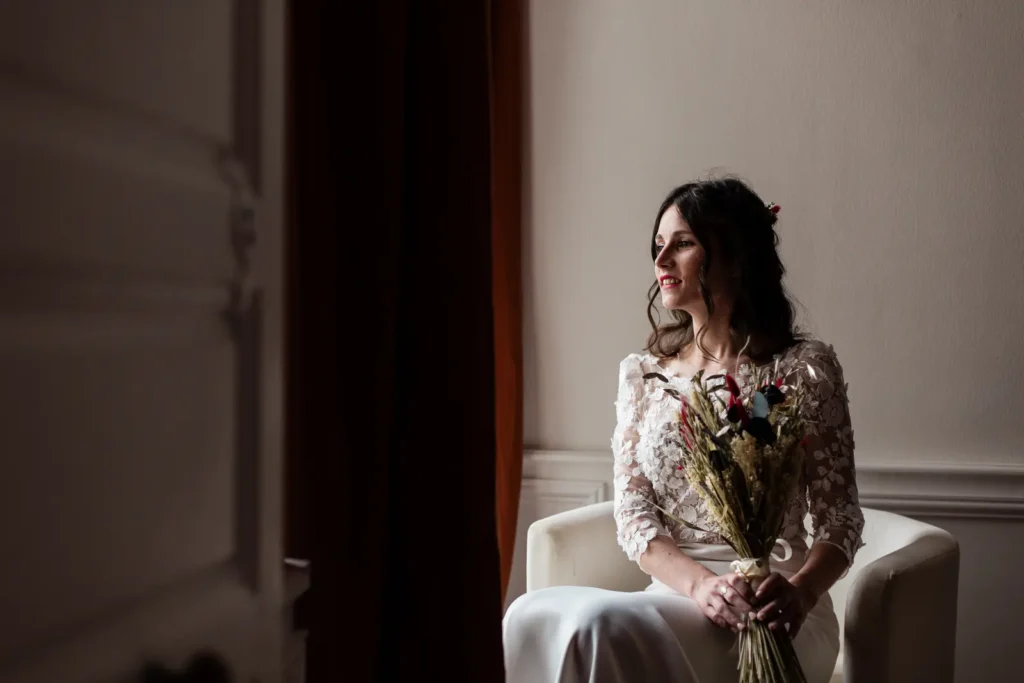 Portrait de la mariée assise avec son bouquet de fleurs séchées, éclairée par une lumière douce près d'une fenêtre au château de la Jahotière.
