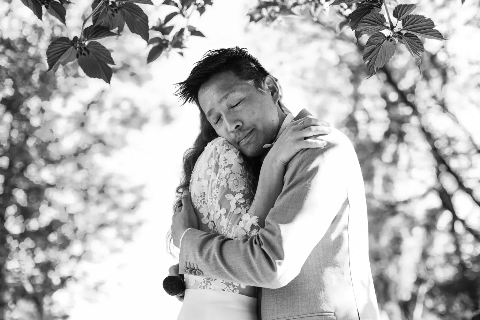 Photo de mariage en noir et blanc capturant une étreinte émouvante entre les mariés sous le feuillage d'un arbre lors de la cérémonie.