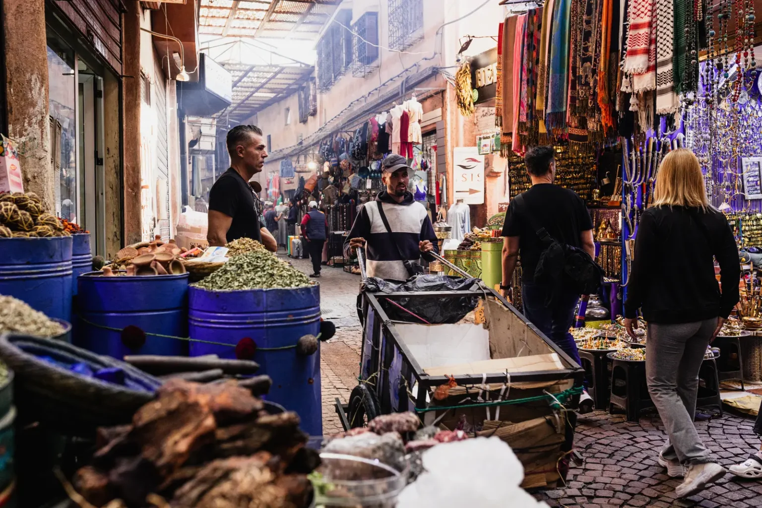 Scène de vie dans les souks de Marrakech : passants et étals colorés de tissus et d'épices lors d'un reportage.