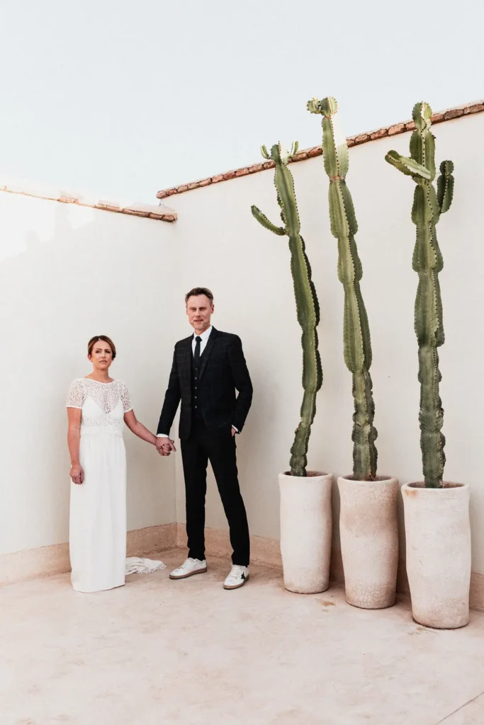 Séance photo de couple de mariés devant un mur blanc et de grands cactus en pots sur une terrasse ensoleillée à Marrakech.
