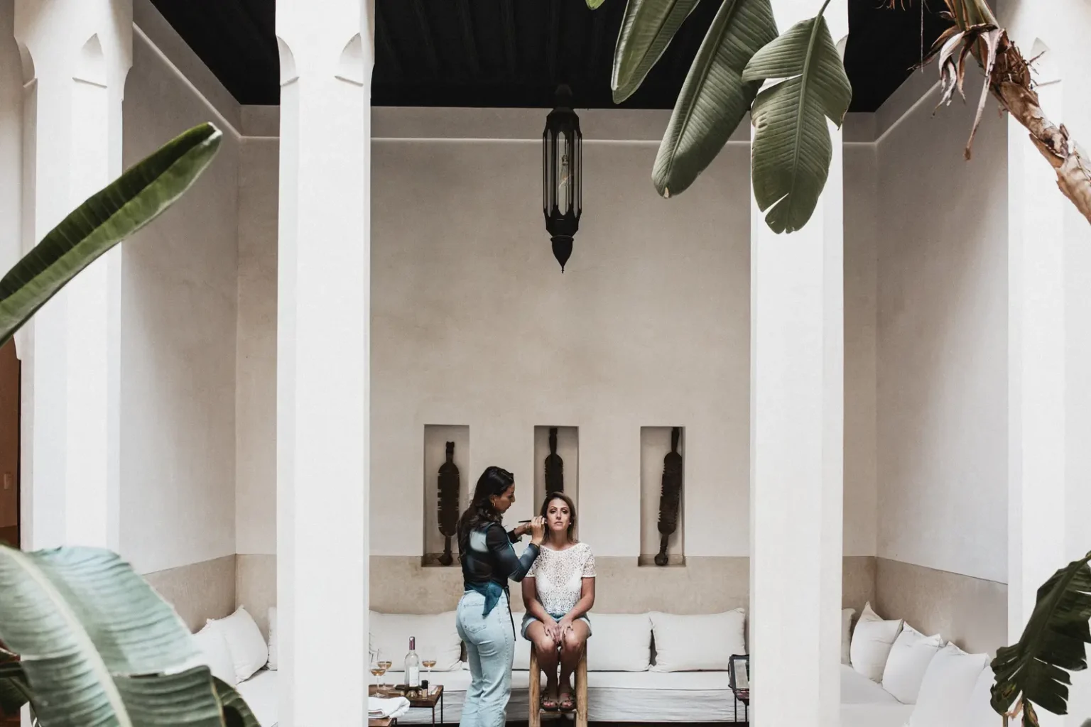 Séance de maquillage de la mariée dans le patio épuré d'un riad à Marrakech, entourée de colonnes blanches et de feuilles de bananier.