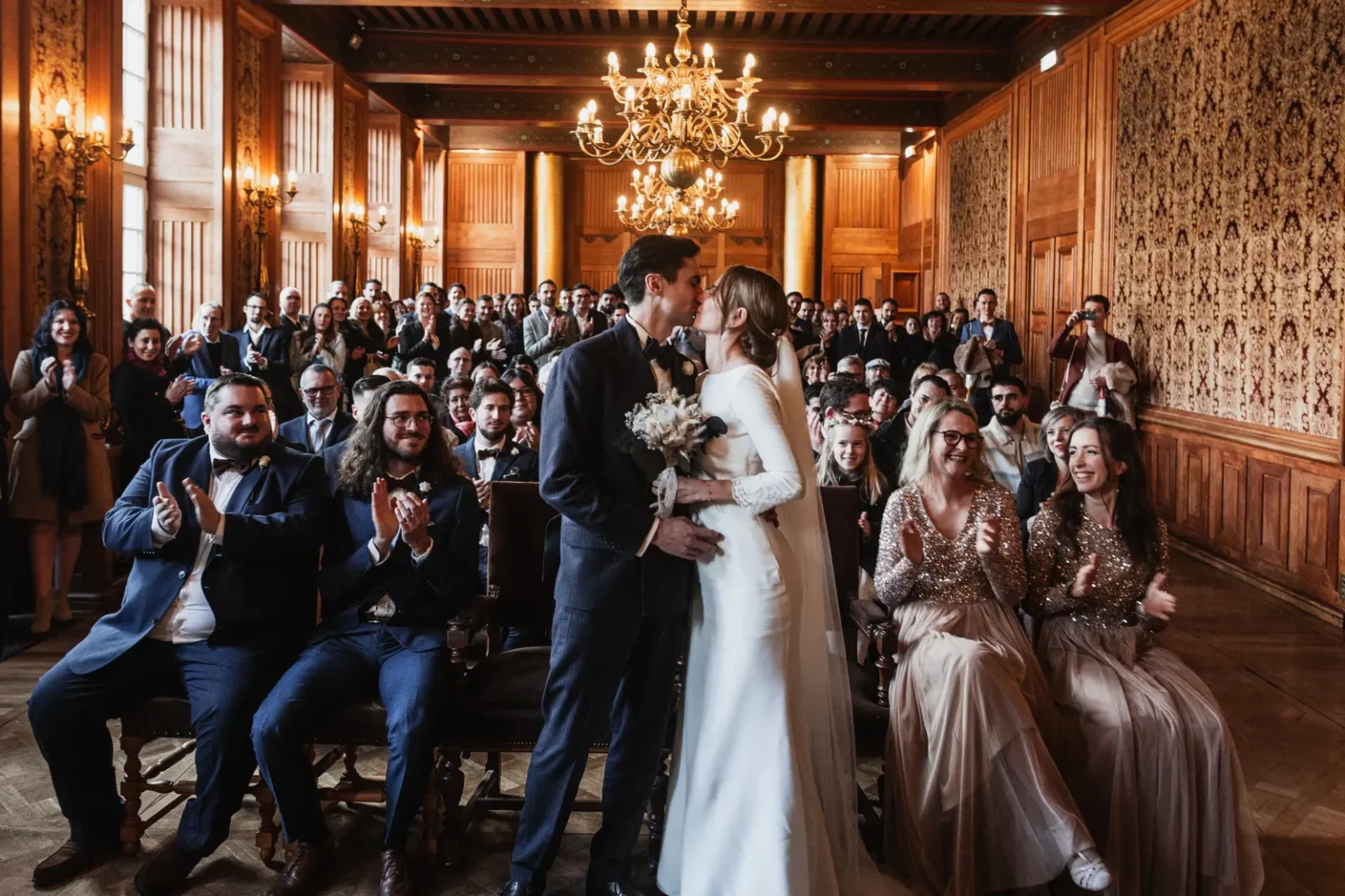 Échange d'un baiser entre les mariés sous les lustres de la salle de cérémonie, entourés de leurs invités qui applaudissent.