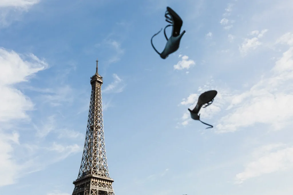 Vue en contre-plongée de la Tour Eiffel à Paris sous un ciel bleu parsemé de nuages, avec une paire de chaussures de mariée à talons hauts lancée en l'air, créant un effet de mouvement dynamique et créatif.