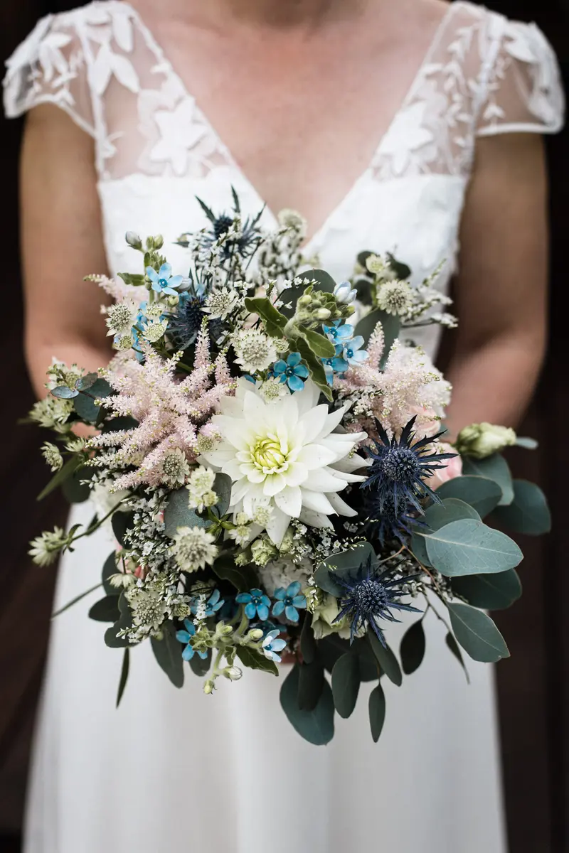 Gros plan sur un bouquet de mariée sauvage et champêtre composé de dahlias blancs, de chardons bleus, d'eucalyptus et de fleurs sauvages
