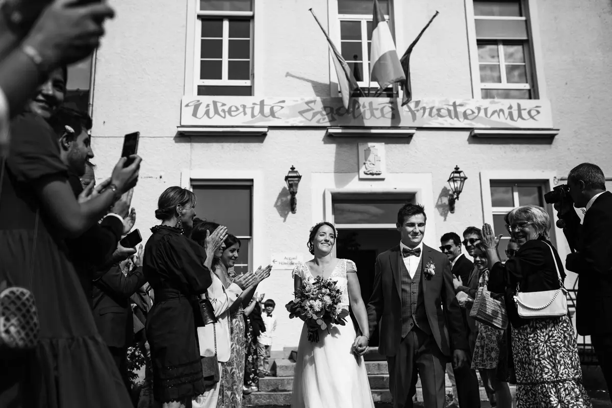 Photo de mariage en noir et blanc montrant la sortie des mariés de la mairie sous les applaudissements des invités, devant la devise "Liberté, Égalité, Fraternité".