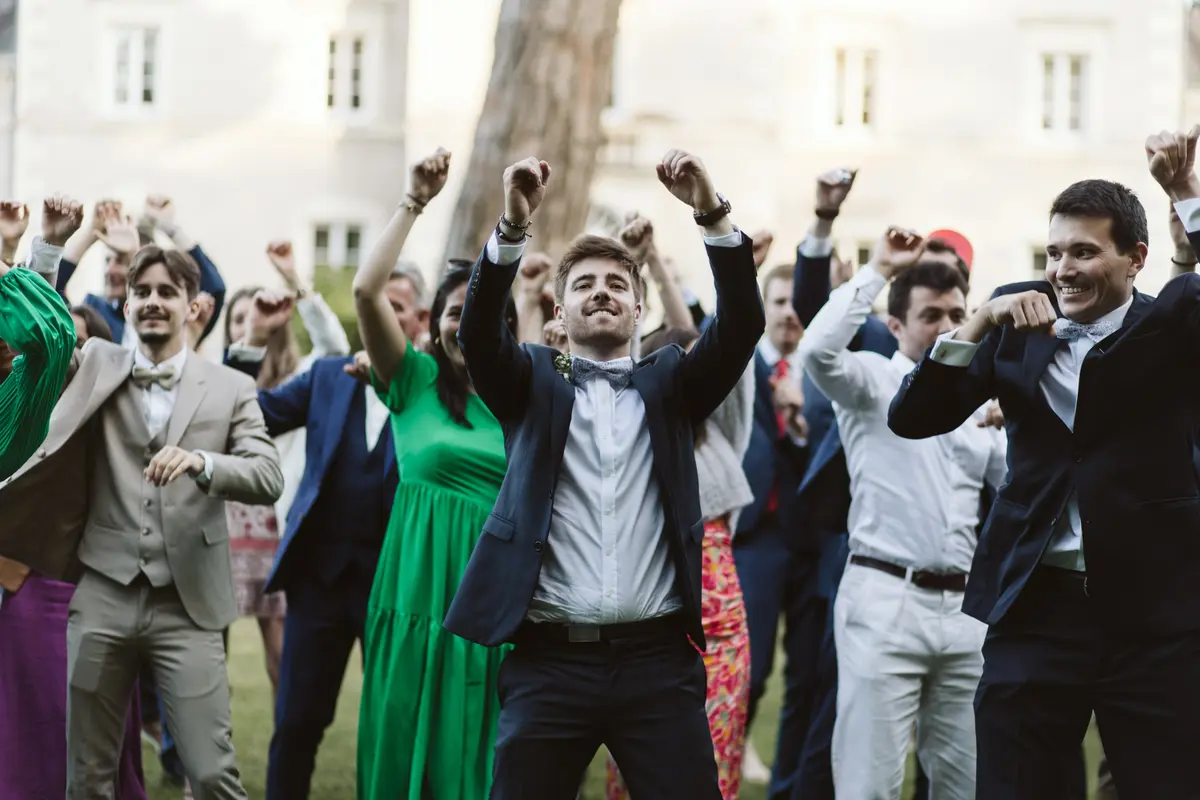 Photo de groupe dynamique lors d'un mariage montrant les invités et le marié les bras levés en train de danser et de célébrer en extérieur.
