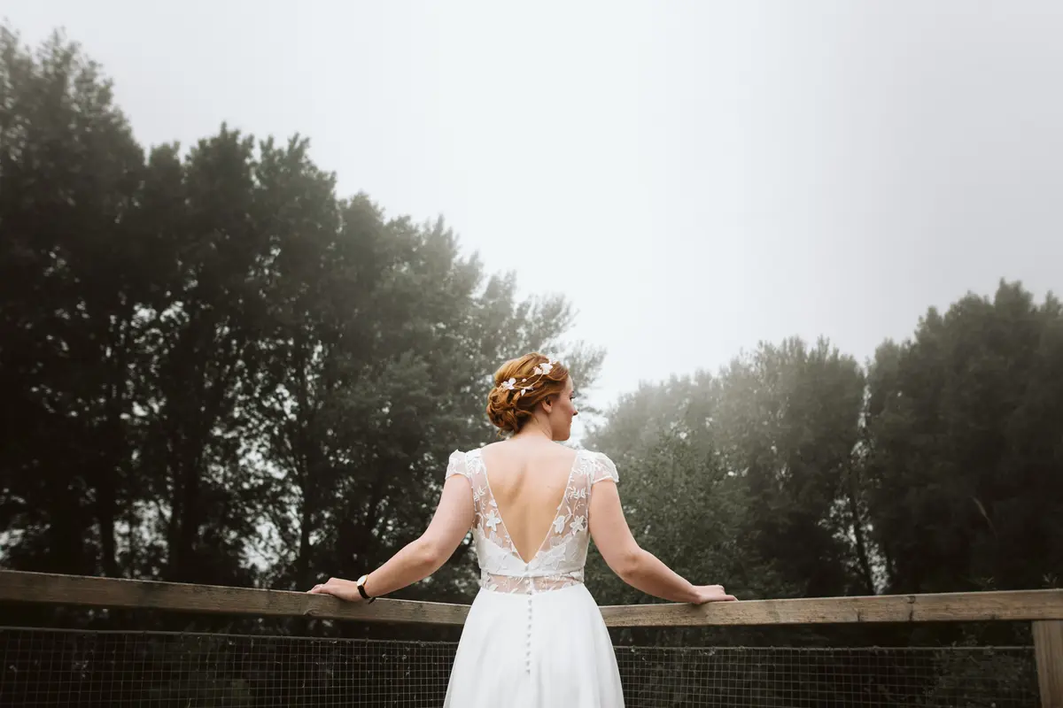 Vue de dos d'une mariée portant une robe avec un décolleté en V en dentelle délicate, regardant vers une forêt brumeuse depuis un balcon en bois.