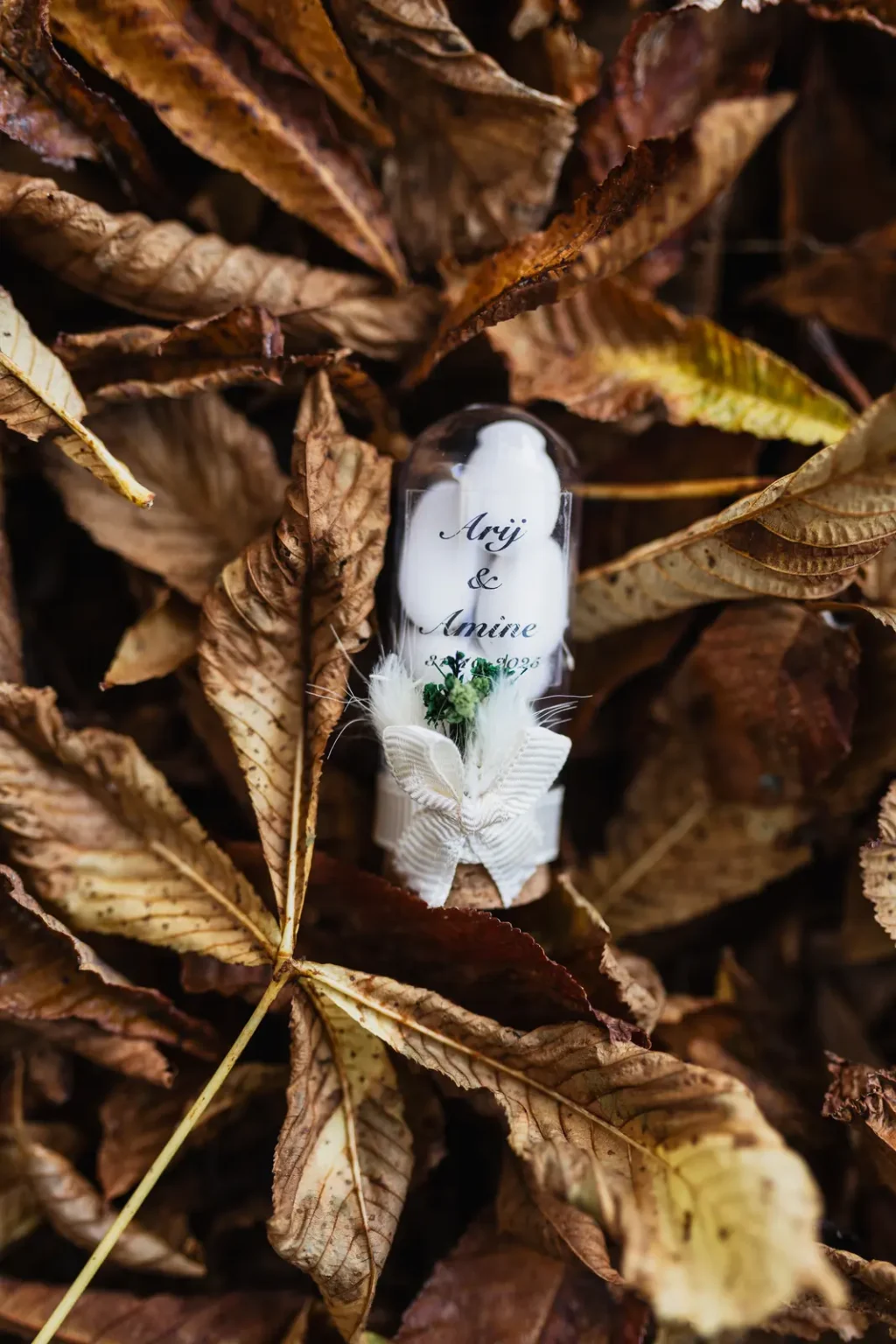 Cadeau d'invités : tube de dragées personnalisé "Arij & Amine" posé sur un lit de feuilles d'automne brunes.