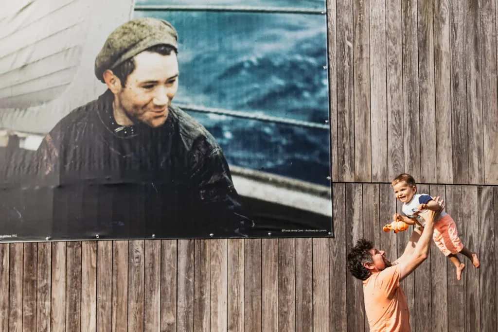 Un papa fair sauter son enfant dans les bras avec une affiche derriere et un marin qui regarde dans leur direction
