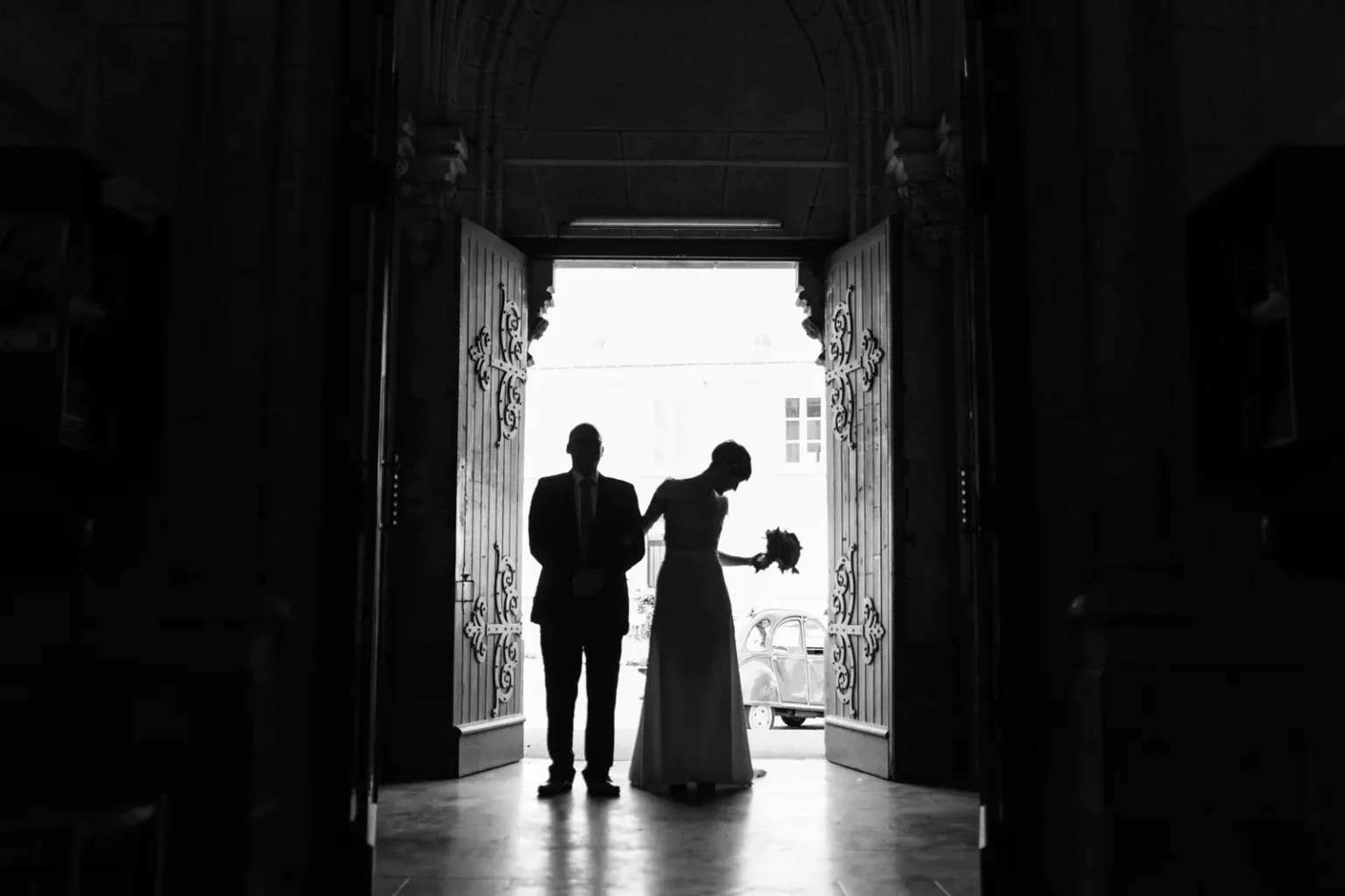 Silhouette en noir et blanc des mariés se tenant côte à côte dans l'encadrement d'un portail d'église orné, une 2CV visible en arrière-plan
