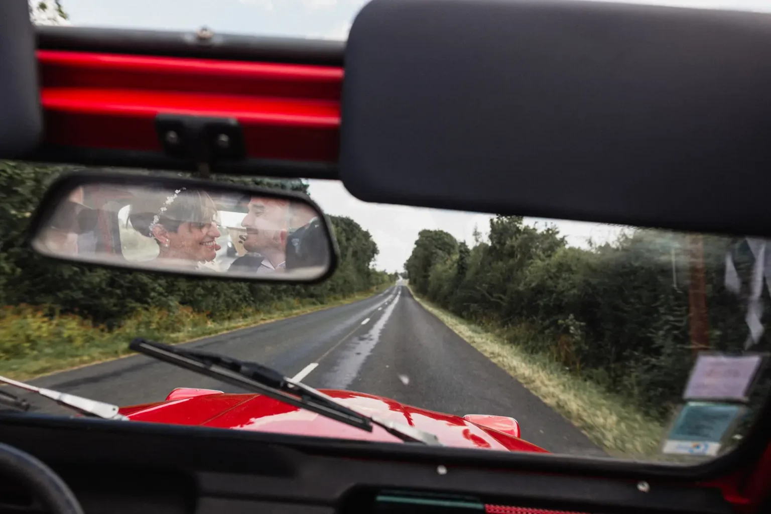 Reflet des mariés dans le rétroviseur d'une 2cv rouge, souriant l'un vers l'autre sur une route de campagne verdoyante