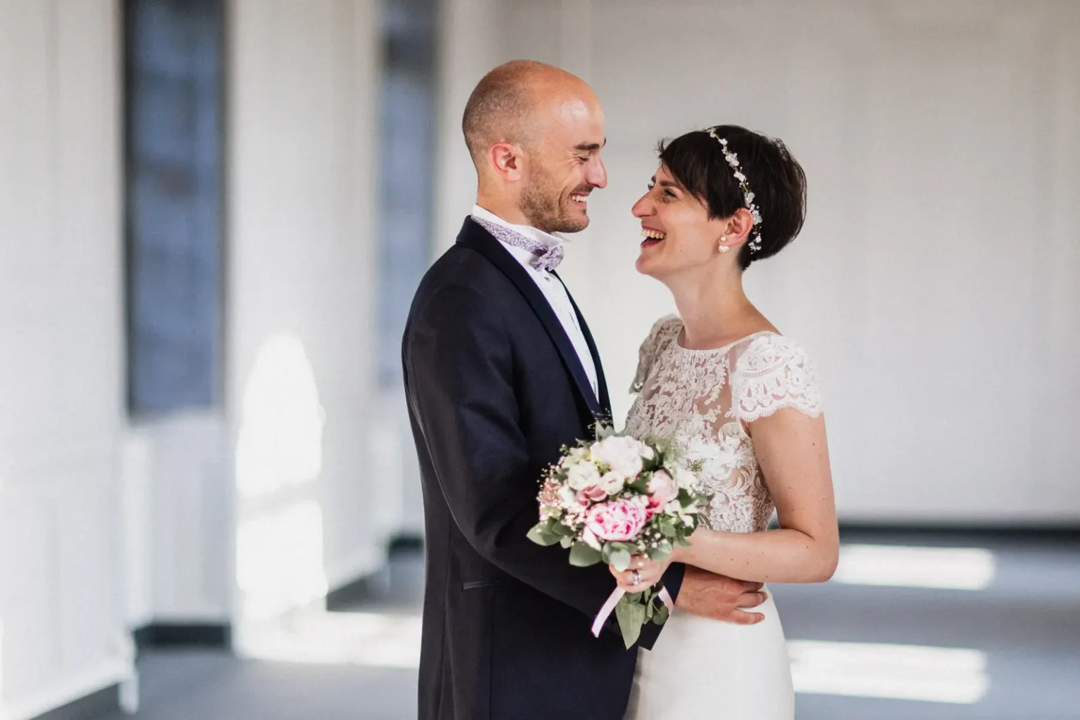 Couple de mariés se regardant en riant, la mariée tenant un bouquet de pivoines roses et blanches, dans une salle lumineuse aux grandes fenêtres