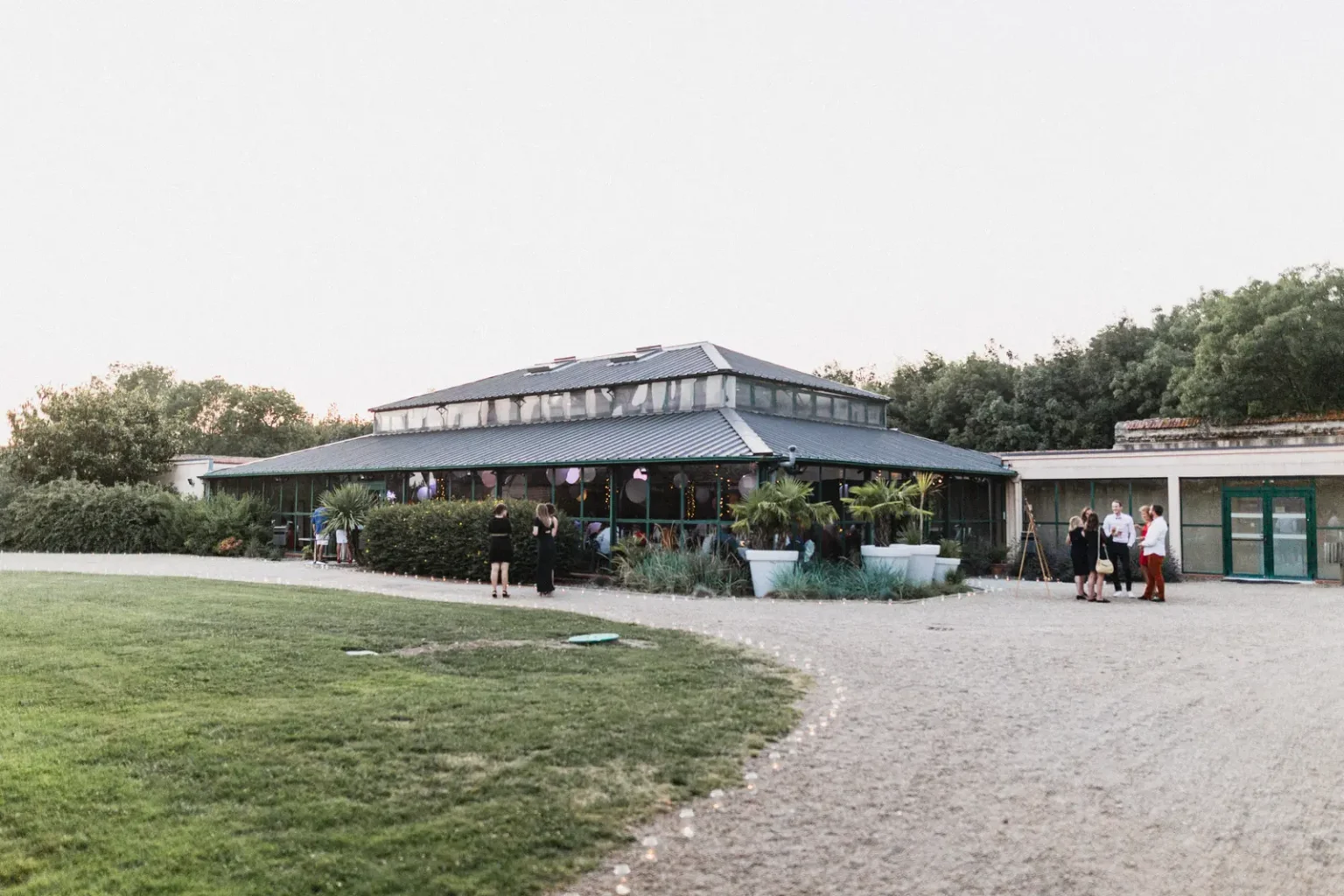 Salle de réception en verre et métal du Domaine des Lys en soirée, entourée de végétation, avec des invités réunis devant l'entrée