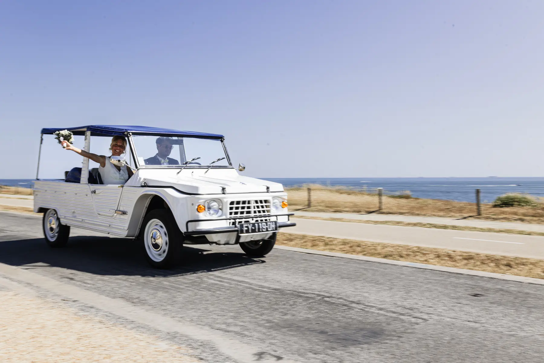 Mariée tenant son bouquet à bord d'une Citroën Méhari blanche conduite par le marié en bord de mer à La Baule.