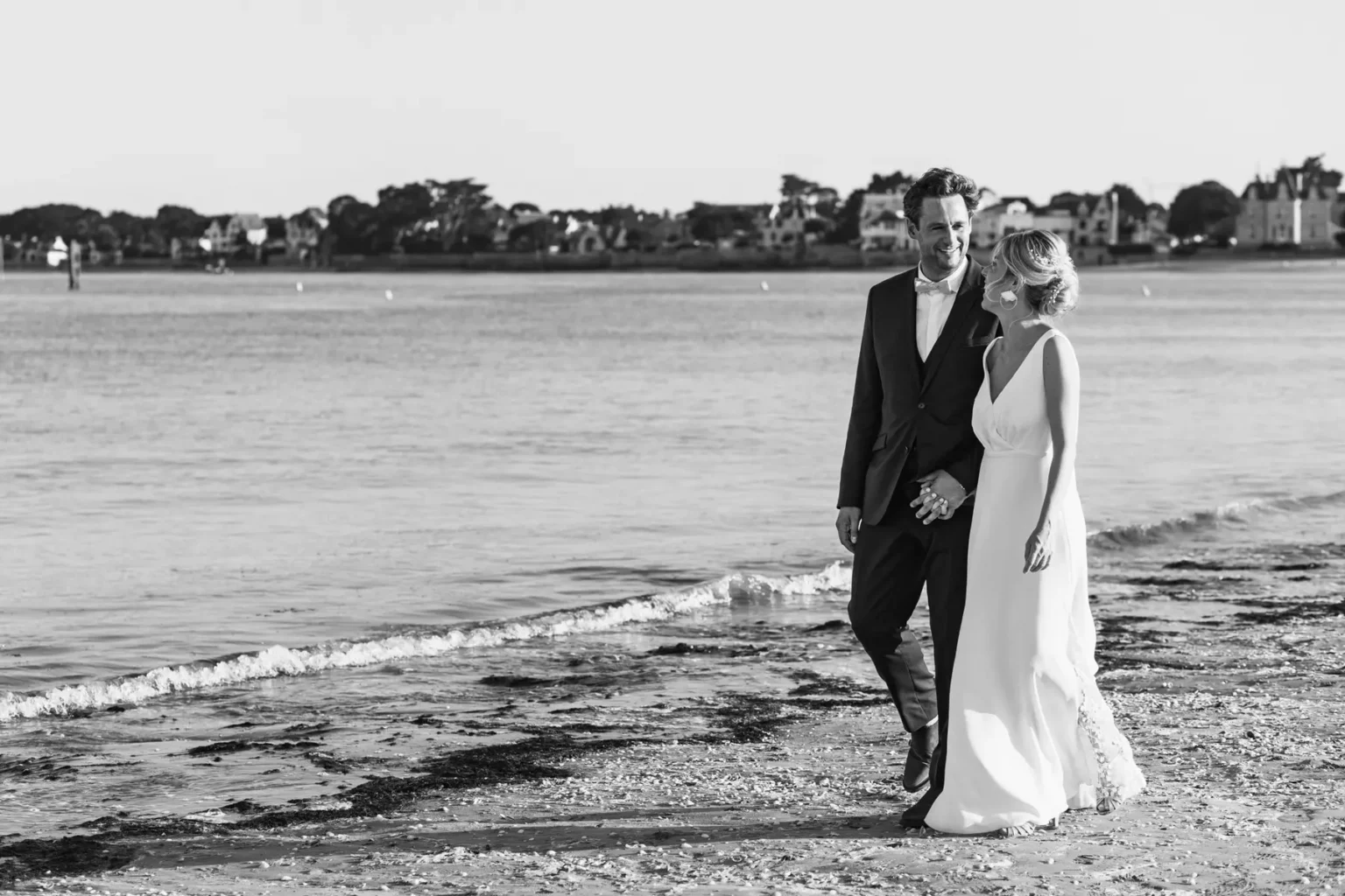 Portrait en noir et blanc des mariés se promenant main dans la main sur le sable au bord de l'eau.