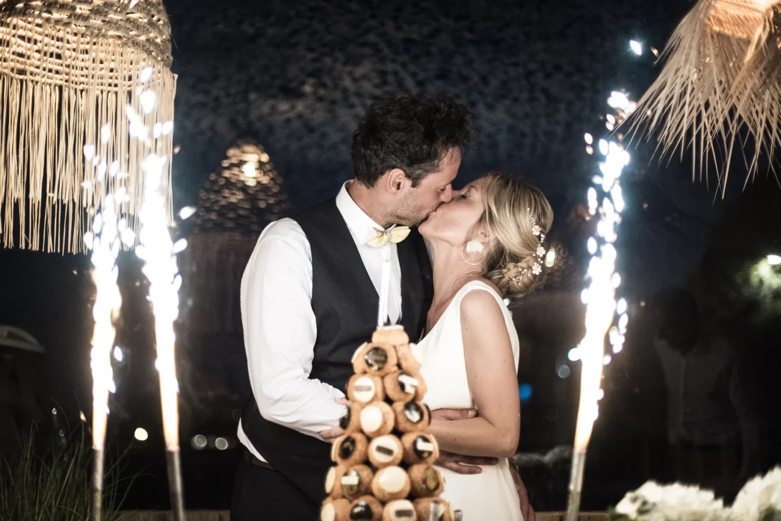Baiser des mariés derrière la pièce montée de choux illuminée par des étincelles lors de la soirée.